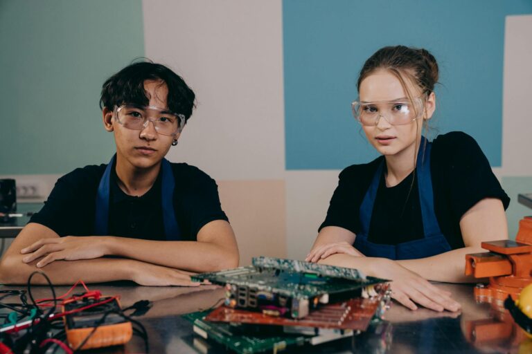 Two young engineers in a workshop with circuit boards, wearing safety glasses.